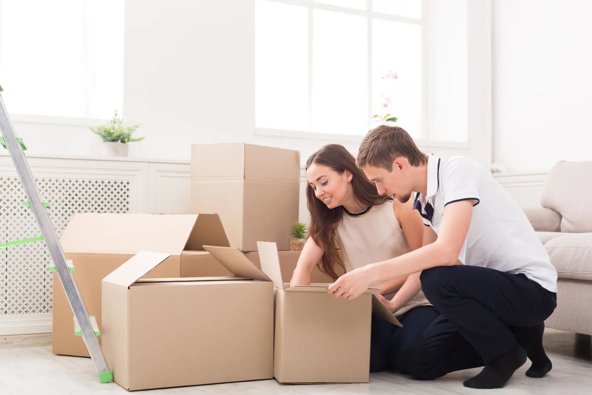 A couple unpacking cardboard boxes in a bright, modern living room, surrounded by greenery and a ladder nearby