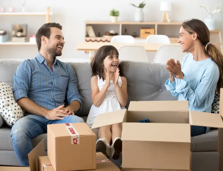A family sits on a couch surrounded by moving boxes, engaging in a joyful conversation, with the child clapping excitedly