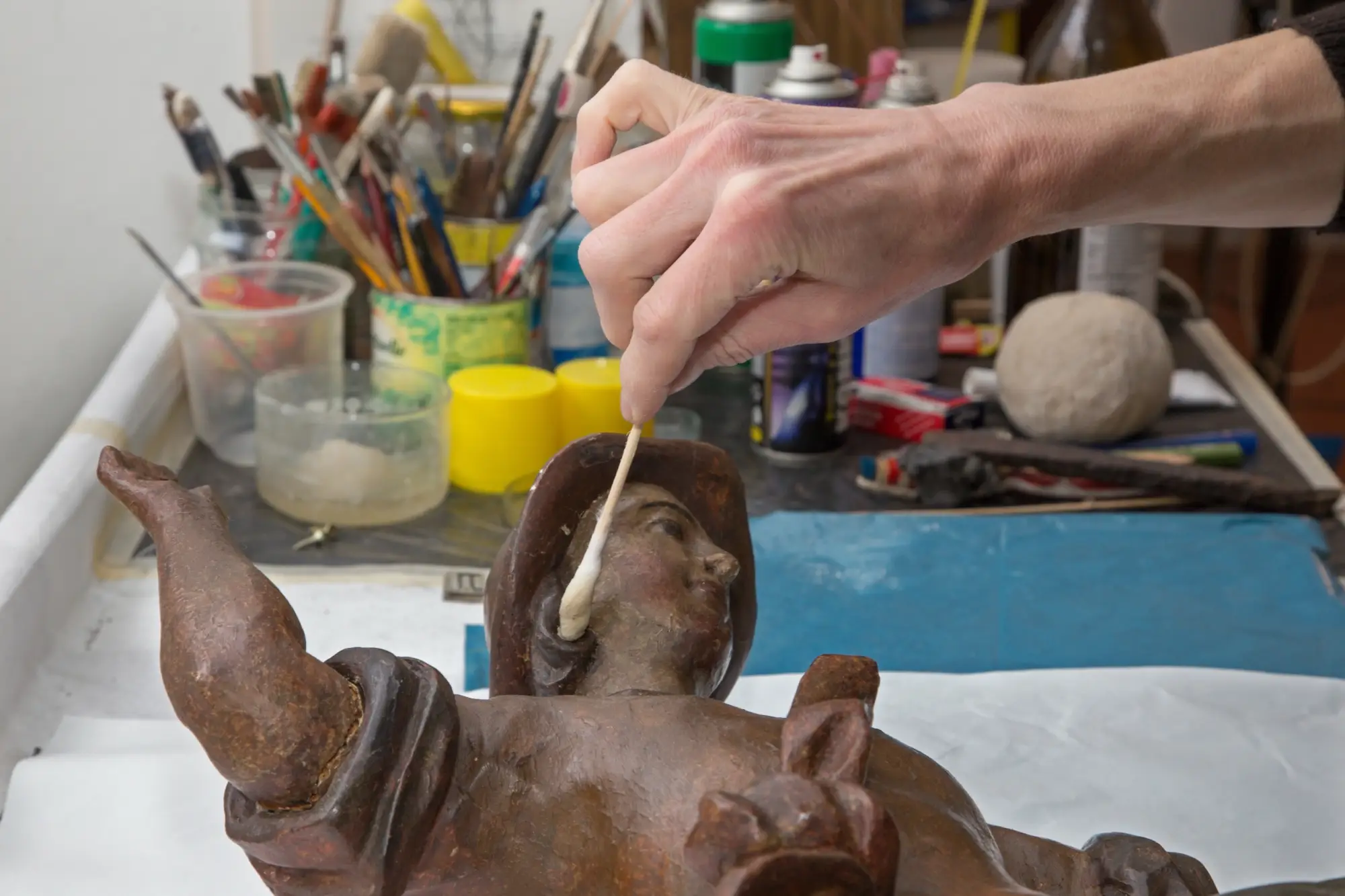 A hand applies finishing touches to a wooden sculpture, surrounded by art supplies and tools on a workbench