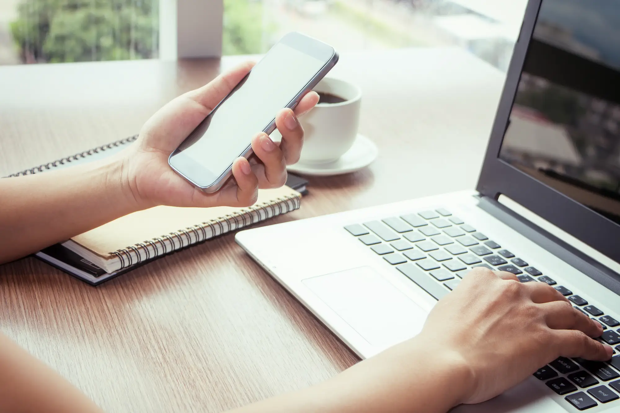 A person holds a smartphone in one hand while typing on a laptop, with a notebook and coffee cup on a wooden table