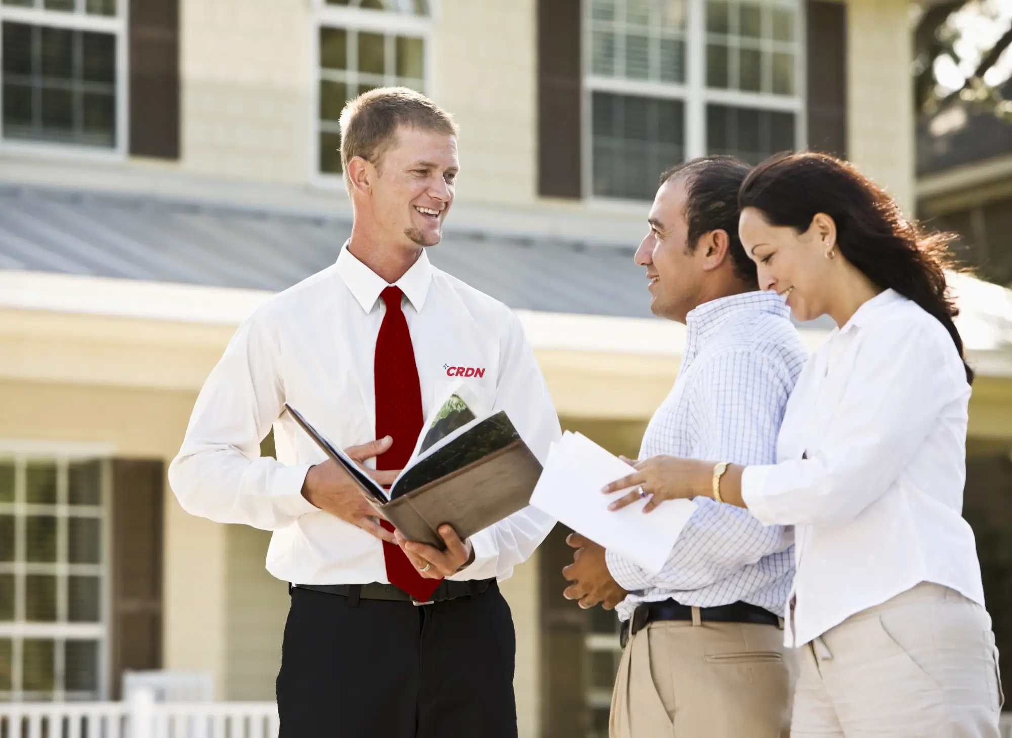 A professional in a white shirt and red tie discusses details with two clients outside a house. Documents and a binder are in their hands