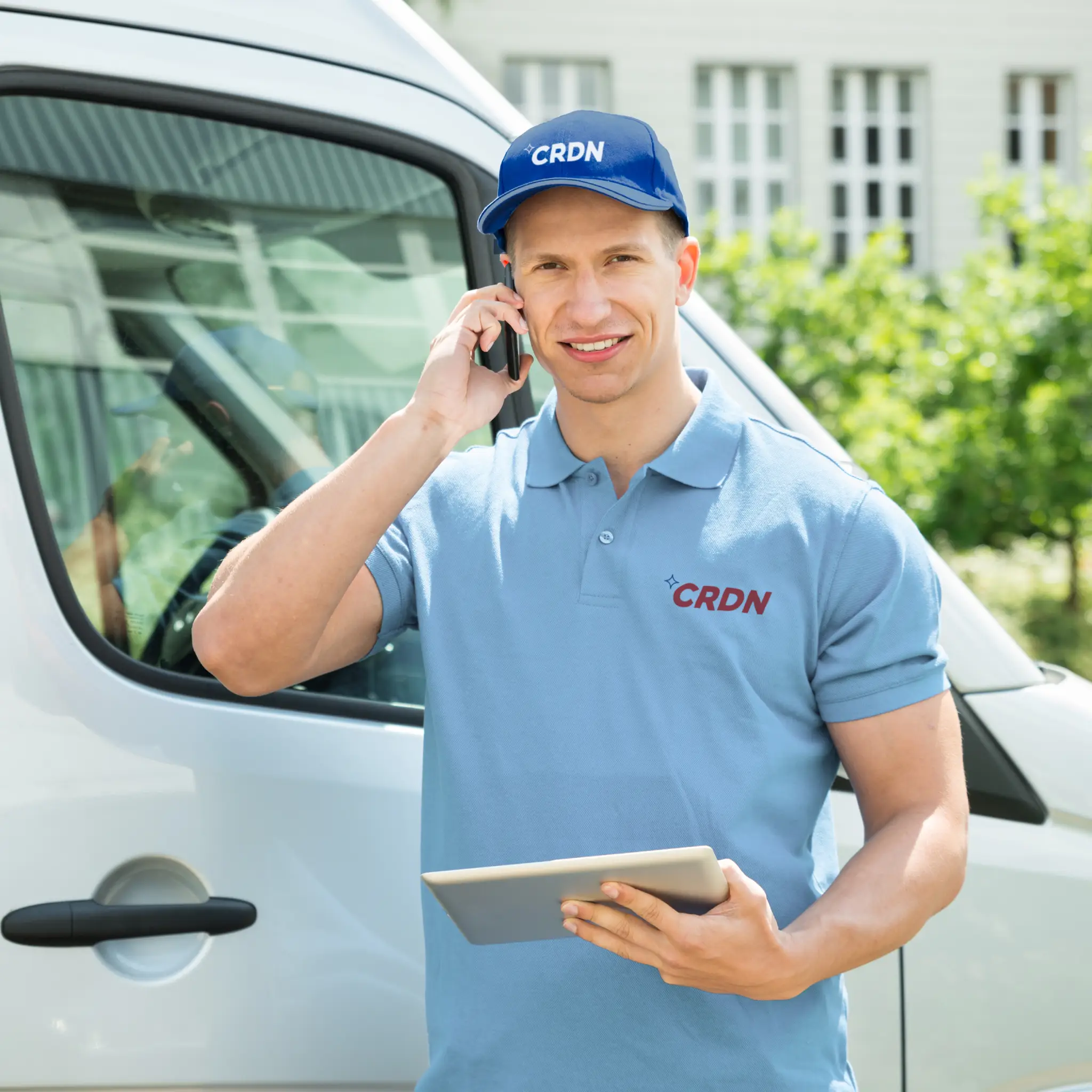 A uniformed worker stands outside a delivery van, holding a tablet and talking on the phone, surrounded by greenery